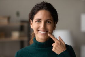 Woman with brown hair in a green sweater smiling and pointing to her teeth